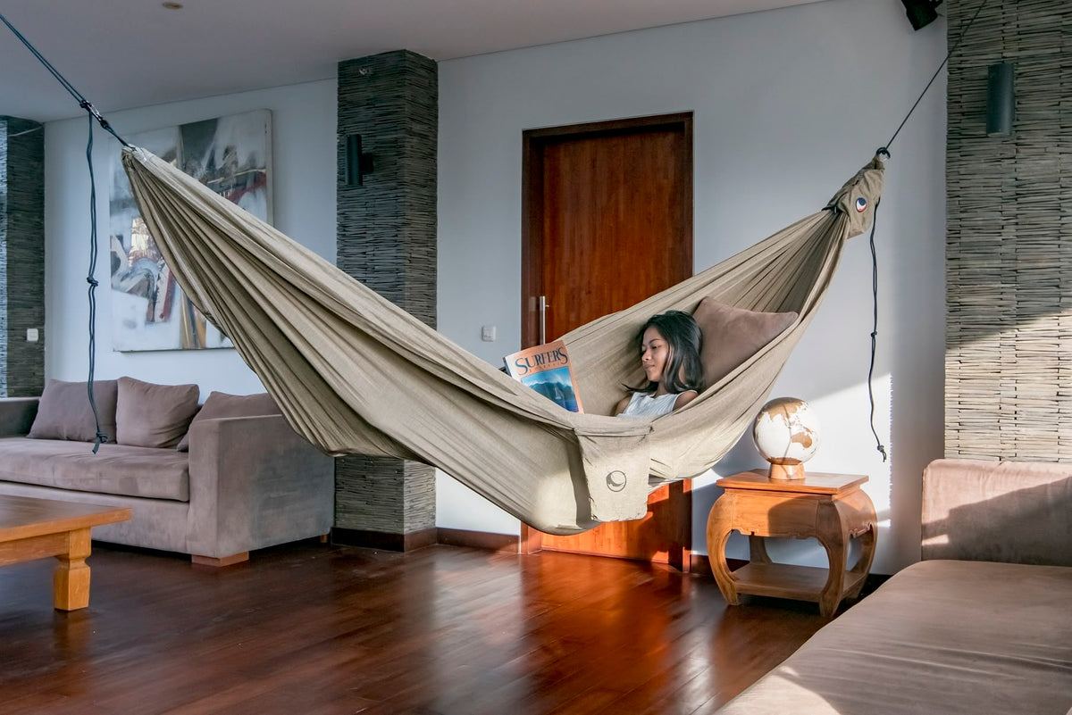 Frau liest ein Surfmagazin in einer Home Hammock 420 in Natural Beige, quer durchs Wohnzimmer gespannt