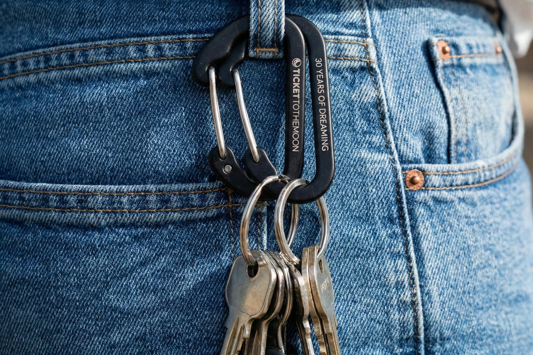 Nahaufnahme von zwei schwarzen Ticket to the Moon Karabinern, die an einer Jeans-Gürtelschlaufe befestigt sind. Einer der Karabiner trägt die Gravur „30 YEARS OF DREAMING“ und hält einen Schlüsselbund.