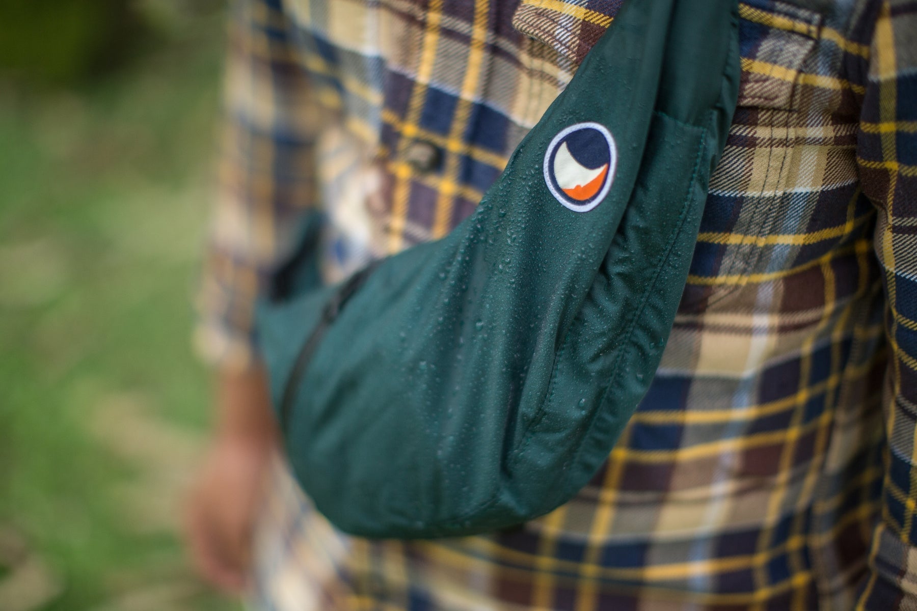 Nahaufnahme einer dunkelgrünen Sling Bag von Ticket to the Moon, die über einem karierten Hemd getragen wird. Wassertropfen perlen auf dem wetterfesten Material ab und demonstrieren die wasserabweisende Funktion der Tasche im Outdoor-Einsatz.