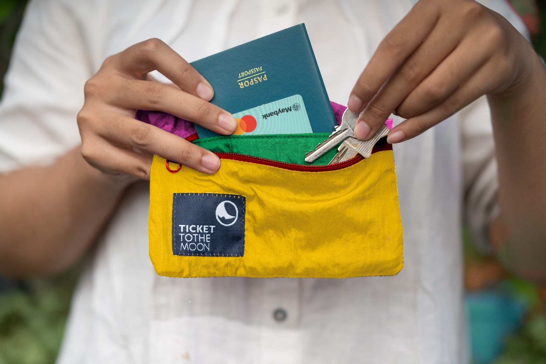 Nahaufnahme einer Person, die einen Reisepass, eine Bankkarte und Schlüssel in eine gelbe Ticket to the Moon Travel Pouch steckt. Das Bild verdeutlicht die kompakte Größe und die praktische Aufteilung des robusten Fallschirmstoffs.