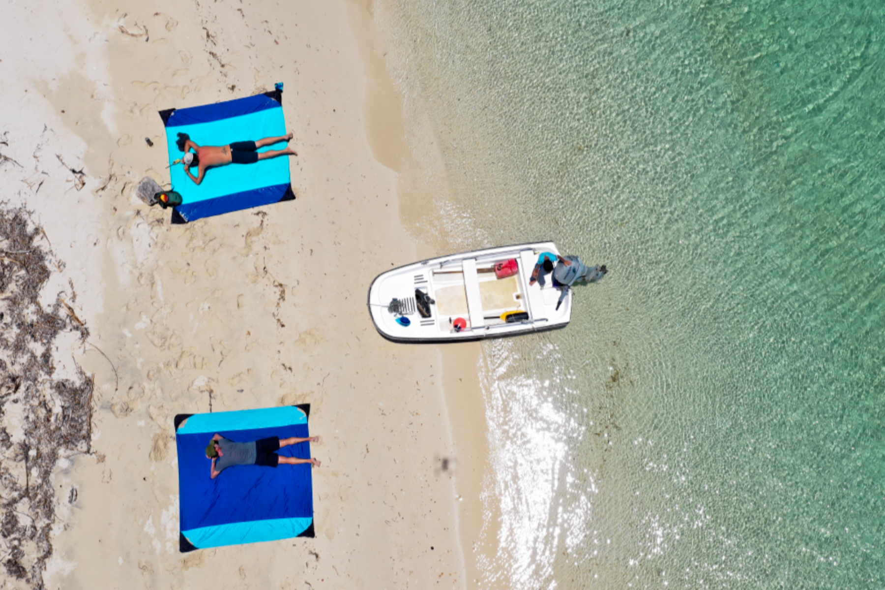 Drohnenaufnahme von zwei Personen, die auf ausgebreiteten Stranddecken am Sandstrand liegen, mit einem Boot im seichten, türkisfarbenen Wasser daneben.
