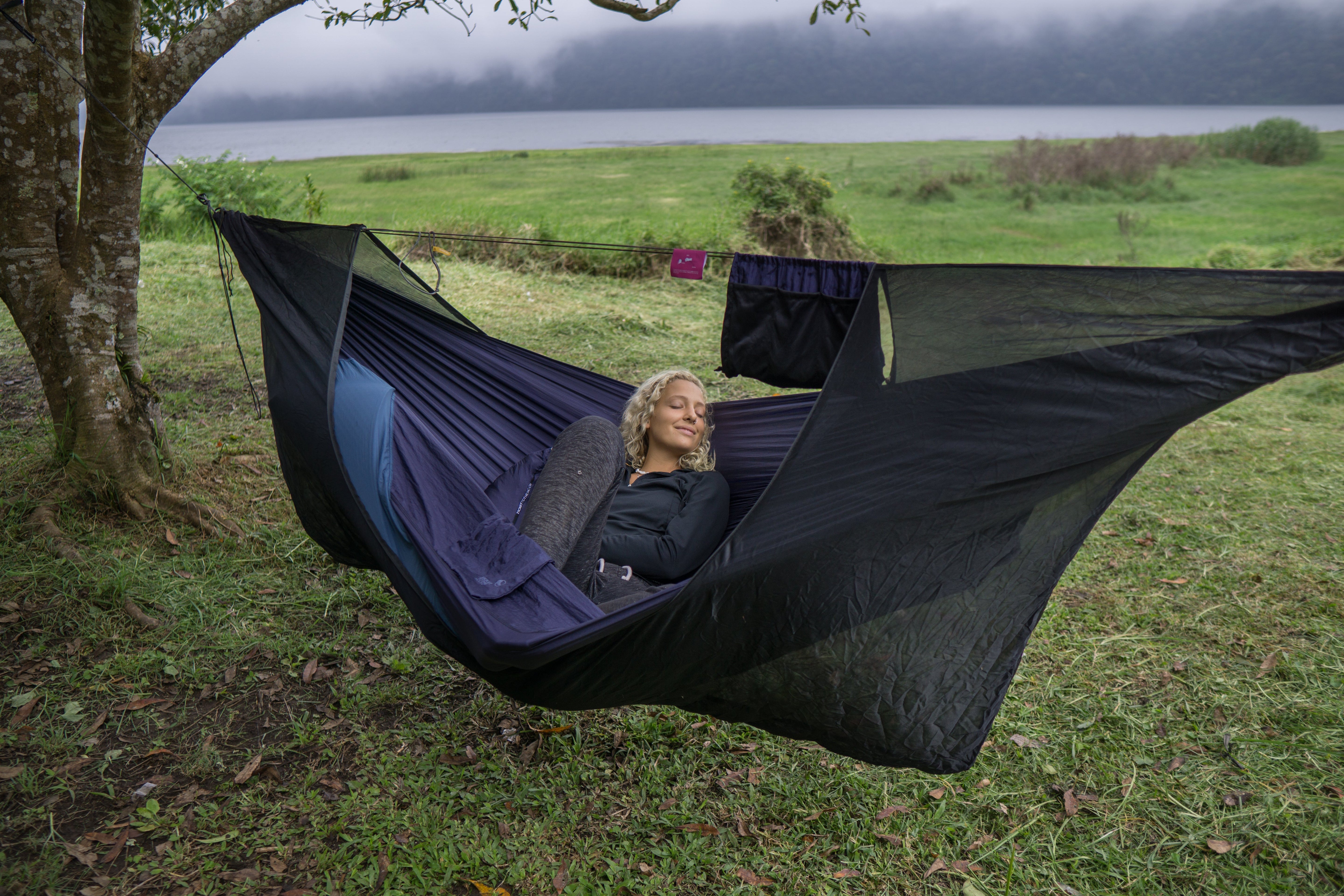 Frau liegt entspannt in einer Hängematte mit aufgespanntem 360° Mückennetz, im Hintergrund eine grüne Wiese, Bäume und ein Seeufer in nebliger Landschaft