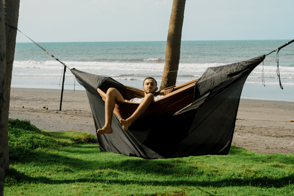 Mann liegt entspannt in einer Hängematte mit schwarzem 360° Mückennetz zwischen Palmen, im Hintergrund Strand und Wellen des Meeres