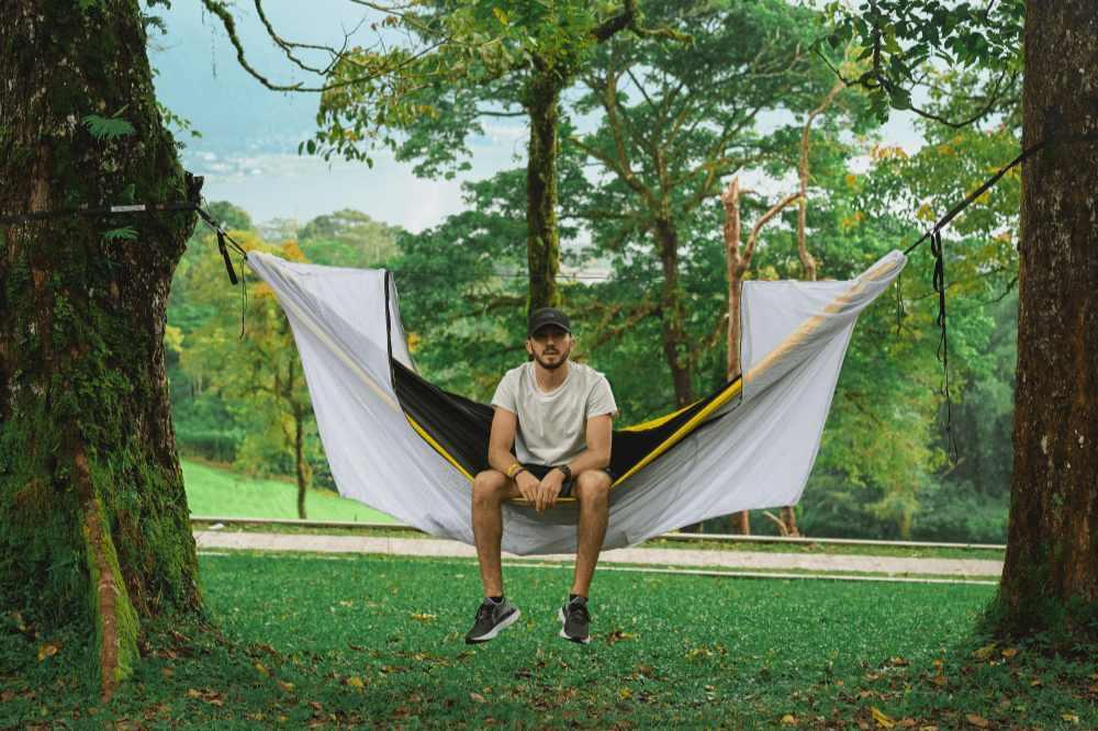 Mann sitzt in einer Hängematte mit geöffnetem weißen 360° Mückennetz, gespannt zwischen zwei Bäumen in grüner Waldlandschaft mit Ausblick ins Tal