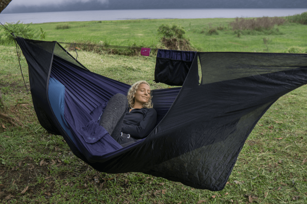 Frau liegt entspannt in einer dunkelblauen Hängematte mit integriertem Moskitonetz und Underquilt, auf einer Wiese mit Seeblick im Hintergrund – ideal für geschützten Outdoor-Schlafkomfort.