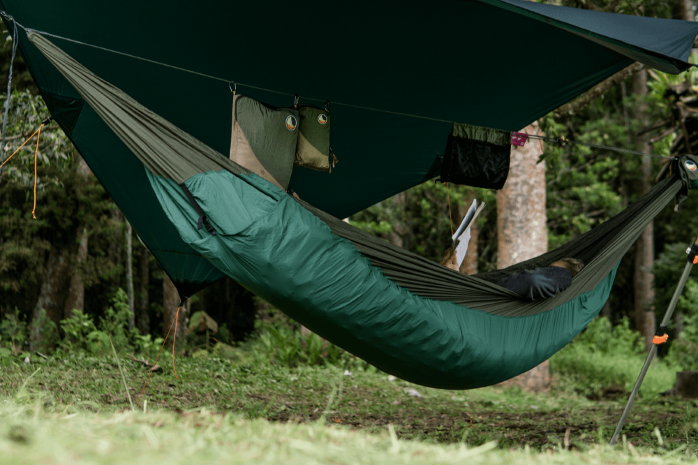 MatOutdoor-Campingszene mit grün-brauner Hängematte, passendem Underquilt und großem Tarp darüber, darunter eine Person mit Buch – umgeben von Wald und aufgehängtem Equipment. Hammock - Ticket To The Moon