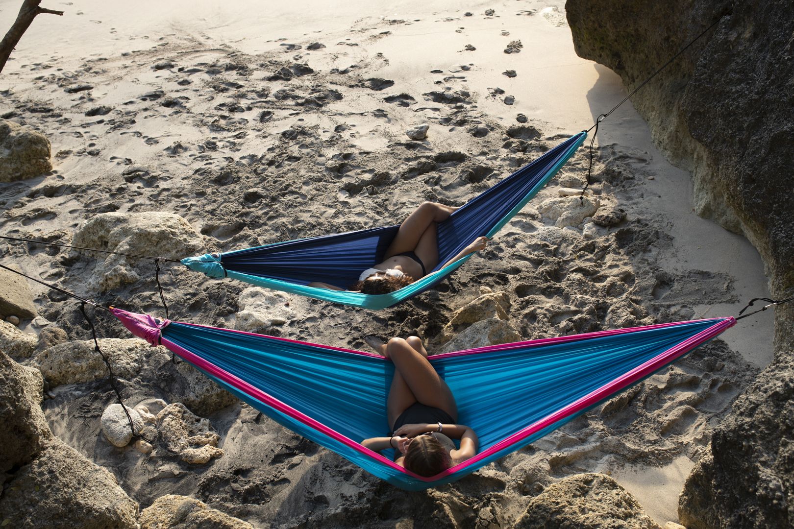 Zwei Personen entspannen in bunten Ticket to the Moon Hängematten zwischen Felsen an einem abgelegenen Sandstrand – Sinnbild für Ruhe, Naturverbundenheit und Freiheit.