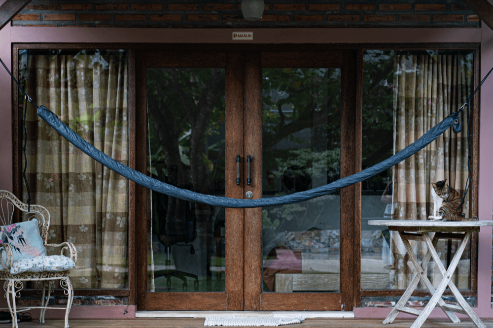 Blau verpackte Hängematte in Hammock Sleeve auf einer Veranda zwischen zwei Befestigungspunkten aufgehängt, im Hintergrund Glastüren und eine sitzende Katze auf einem Tisch