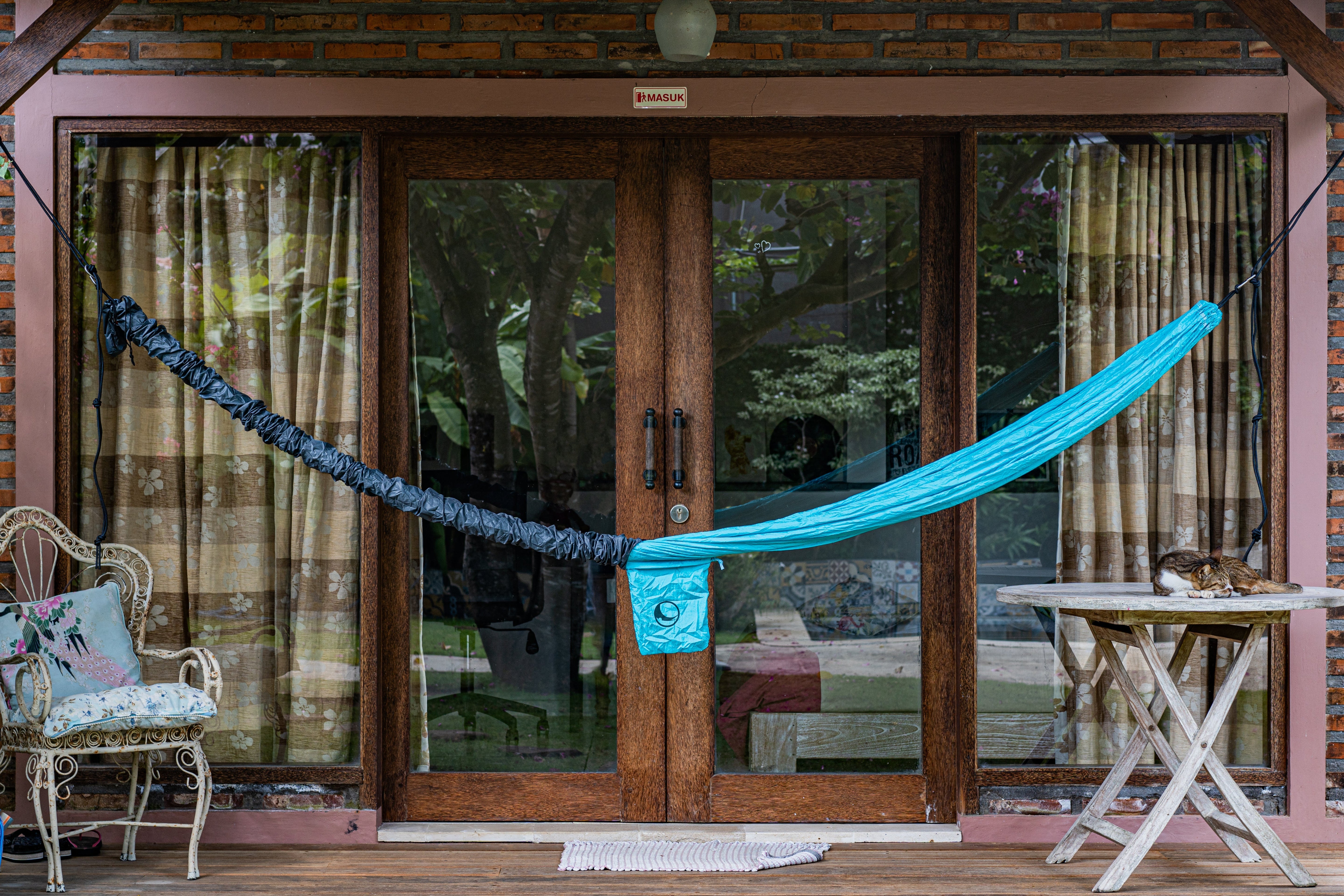 Teilweise geöffnete Hammock Sleeve mit sichtbar aufgehängter türkisfarbener Hängematte auf einer Veranda vor Glastüren, Katze liegt auf einem Tisch