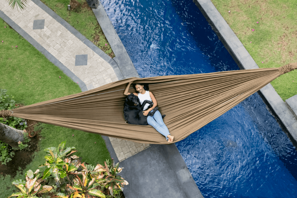 Frau liegt mit schwarzem Hund in einer braunen Ticket to the Moon HOME HAMMOCK, die über einem Pool gespannt ist – von oben fotografiert.
