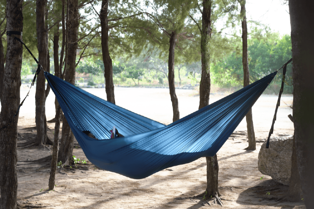 Die LIGHTEST HAMMOCK Hängematte in Blau, aufgehängt zwischen zwei Bäumen, mit Sonnenlicht, das durch das Gewebe scheint, während jemand entspannt darin liegt.