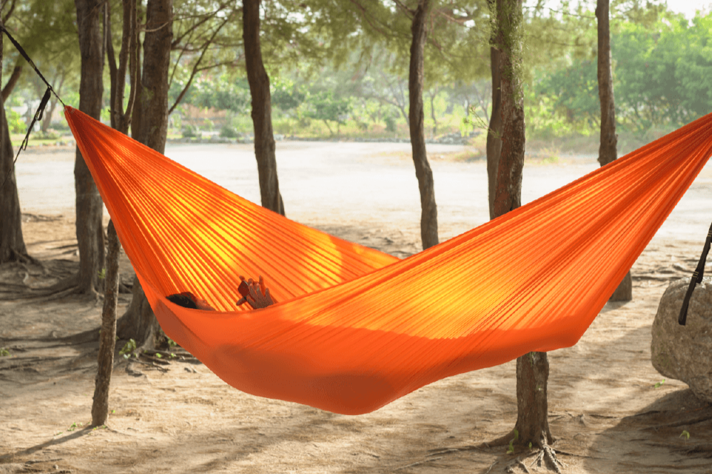 Die LIGHTEST HAMMOCK in Orange, aufgehängt zwischen zwei Bäumen, mit Sonnenlicht, das durch das Gewebe scheint, während jemand entspannt darin liegt.