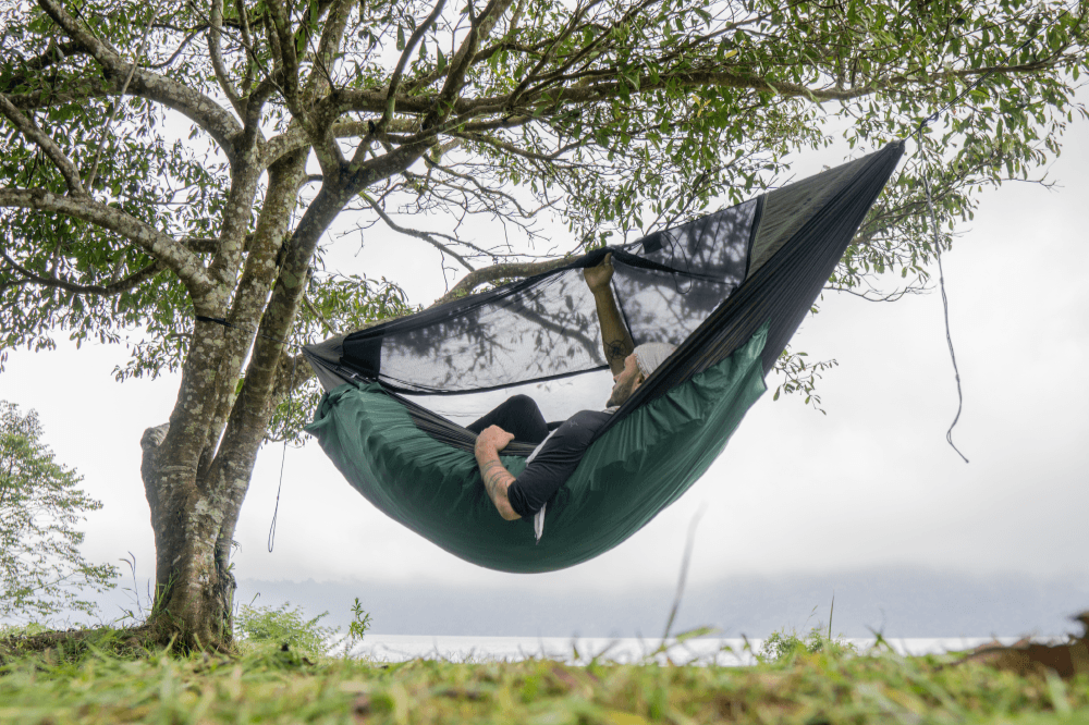 Person liegt in der MAT HAMMOCK mit Moskitonetz und integrierter Matratzenhülle – komfortables Outdoor-Setup unter Bäumen mit Blick auf See oder Berge.