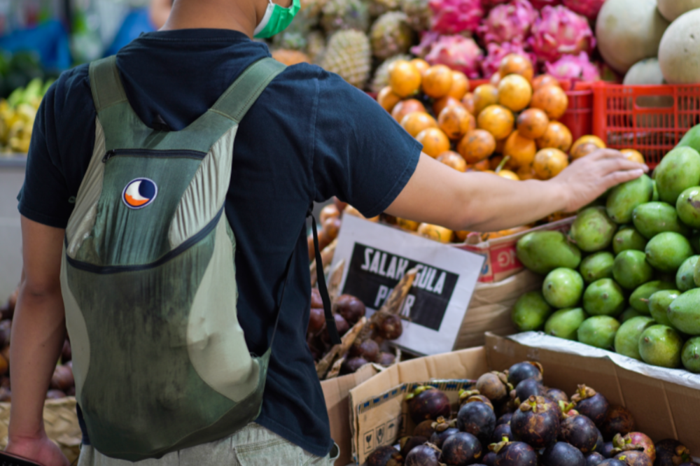 Person mit grünem Mini Backpack von Ticket to the Moon beim Einkauf auf einem tropischen Markt, umgeben von buntem Obst wie Mangos, Mangostanen und Drachenfrüchten.