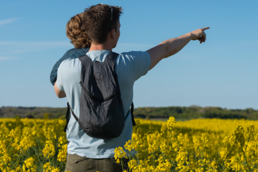Person mit Mini Backpack von Ticket to the Moon auf dem Rücken, trägt ein Kind und zeigt in ein weites Rapsfeld – symbolisiert Leichtigkeit und Outdoor-Abenteuer.