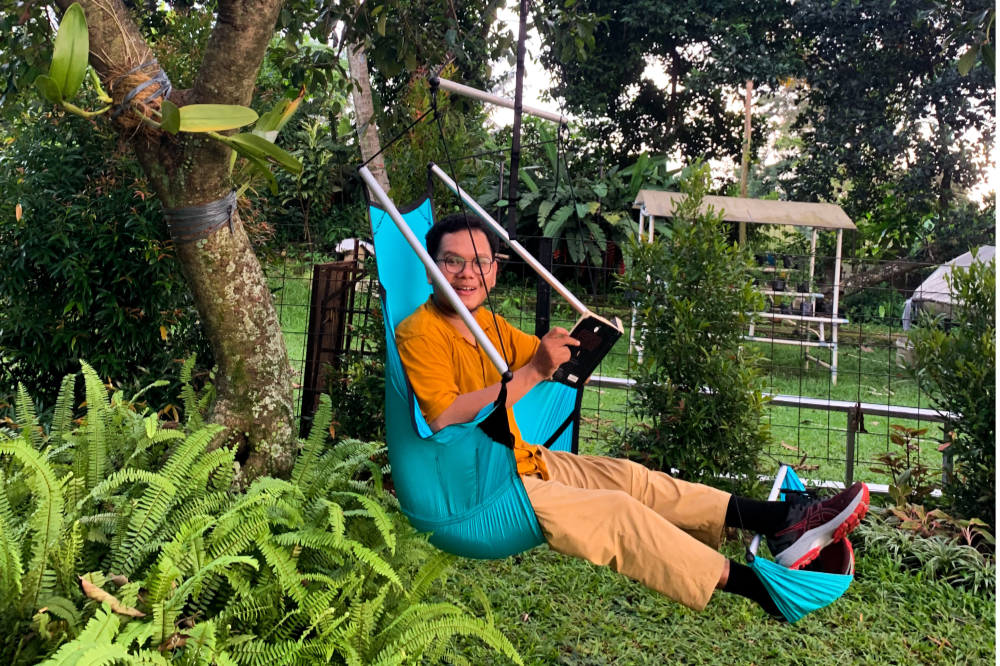 Person sitzt entspannt im türkisfarbenen MOON CHAIR mit Fußstütze im Garten, umgeben von Farnen und tropischen Pflanzen.