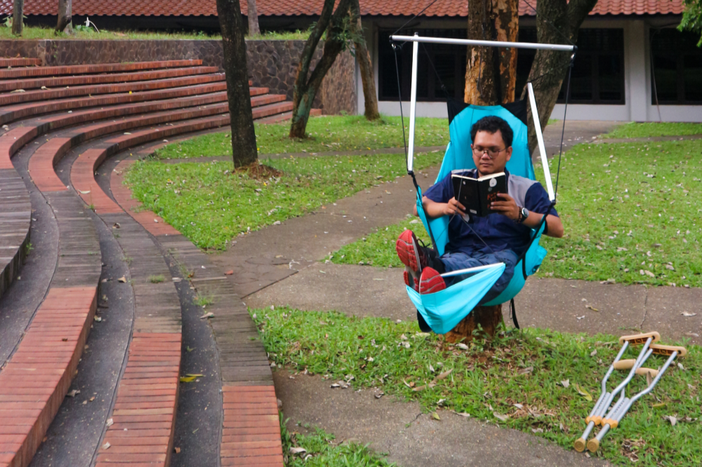 Person sitzt im türkisfarbenen MOON CHAIR und liest in einem Notizbuch, während sie an einem Baum in einem Park aufgehängt ist; Krücken liegen am Boden.