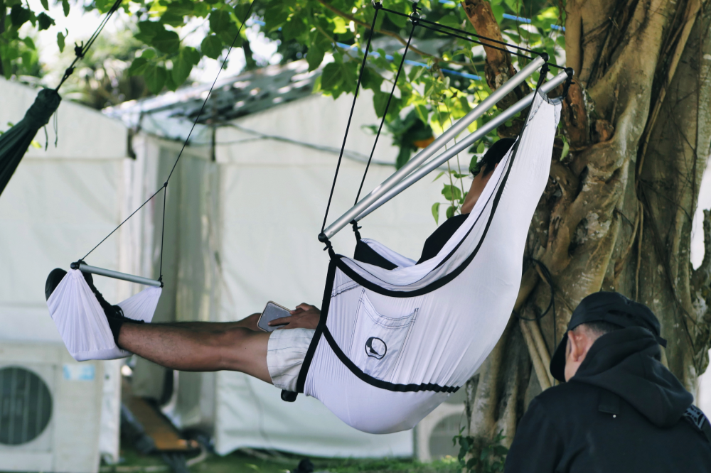 Person sitzt entspannt in einem weißen MOON CHAIR mit Fußstütze, aufgehängt an einem Baum, während jemand danebensteht und zuschaut.