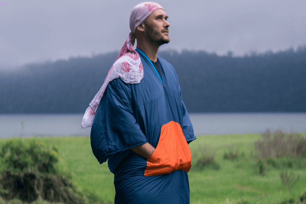 Person trägt die Moonblanket wie einen Mantel mit überkreuzten Händen in der großen Außentasche, steht auf einer grünen Wiese mit Blick auf einen See und bewaldete Hügel im Hintergrund