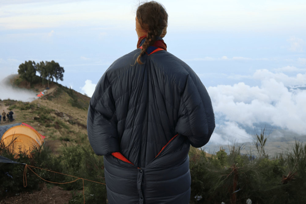 Person mit zusammengebundener Moonblanket von hinten zu sehen, steht auf einem Berg mit Blick über Wolken und Landschaft, im Vordergrund ein Zelt und niedrige Vegetation