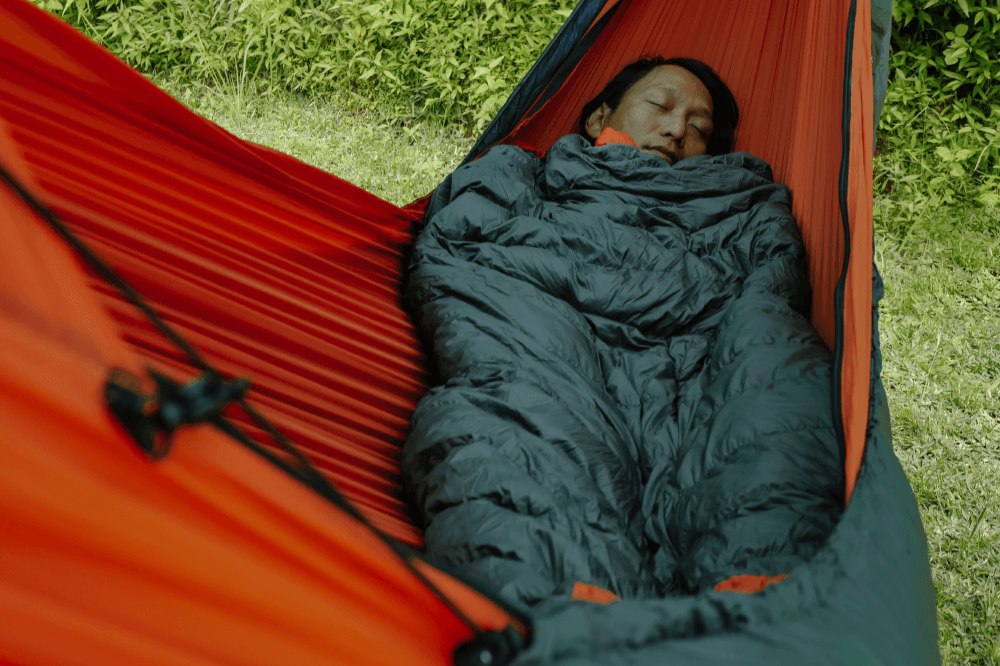 Person liegt schlafend in einer Hängematte, eingewickelt in eine dunkelgraue Moonblanket mit orangen Details, Umgebung ist grün und natürlich