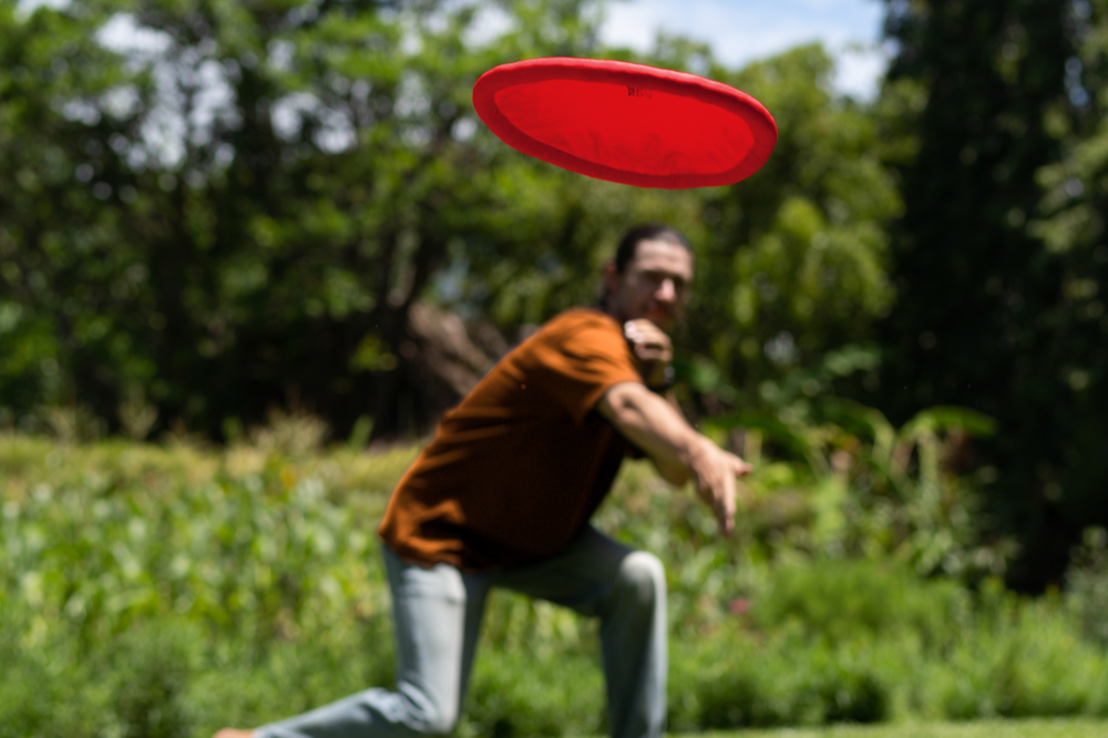 Person wirft eine rote Moondisc auf einer Wiese, die Frisbee schwebt im Vordergrund, Hintergrund unscharf.