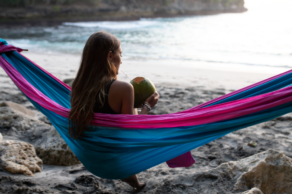 Frau sitzt in einer pink-blauen Hängematte am Strand und trinkt aus einer Kokosnuss.