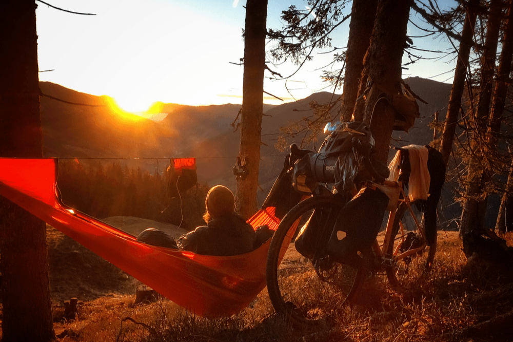 Person liegt in einer PRO HAMMOCK in leuchtendem Orange zwischen Bäumen, mit Fahrrad und Gepäck daneben, Sonnenuntergang über Berglandschaft im Hintergrund.