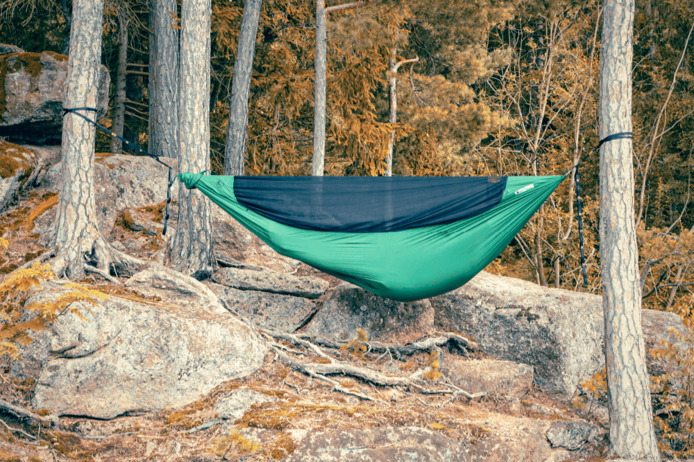 PRO HAMMOCK in grün mit schwarzem Moskitonetz, aufgehängt zwischen Bäumen auf felsigem Untergrund im herbstlich gefärbten Wald.