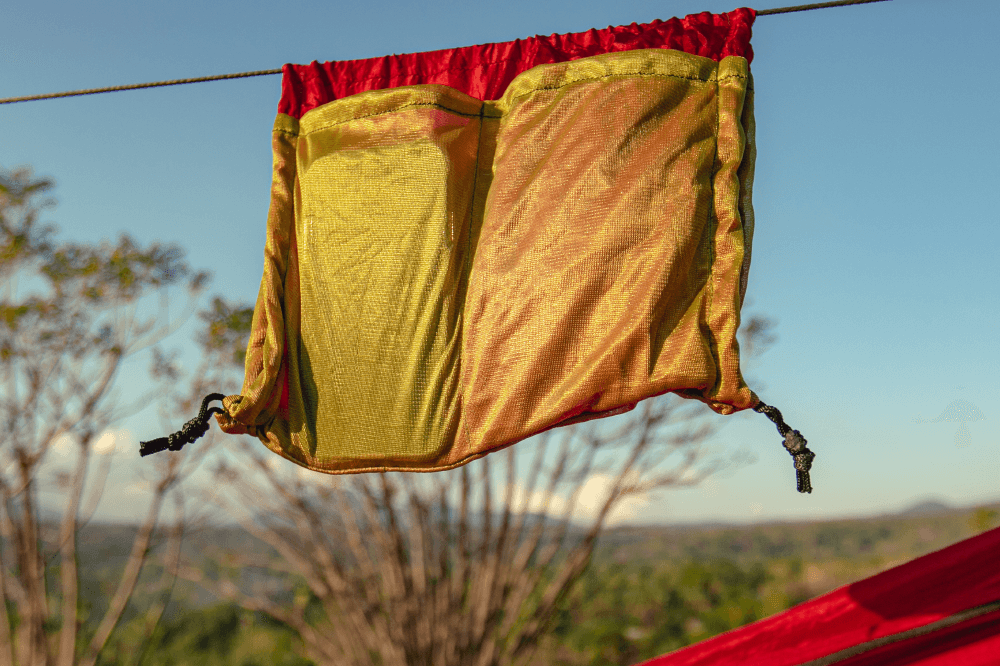 Aufbewahrungstasche der PRO HAMMOCK in Rot-Gelb, aufgehängt an einer Leine mit Hintergrund aus Himmel und Wald.