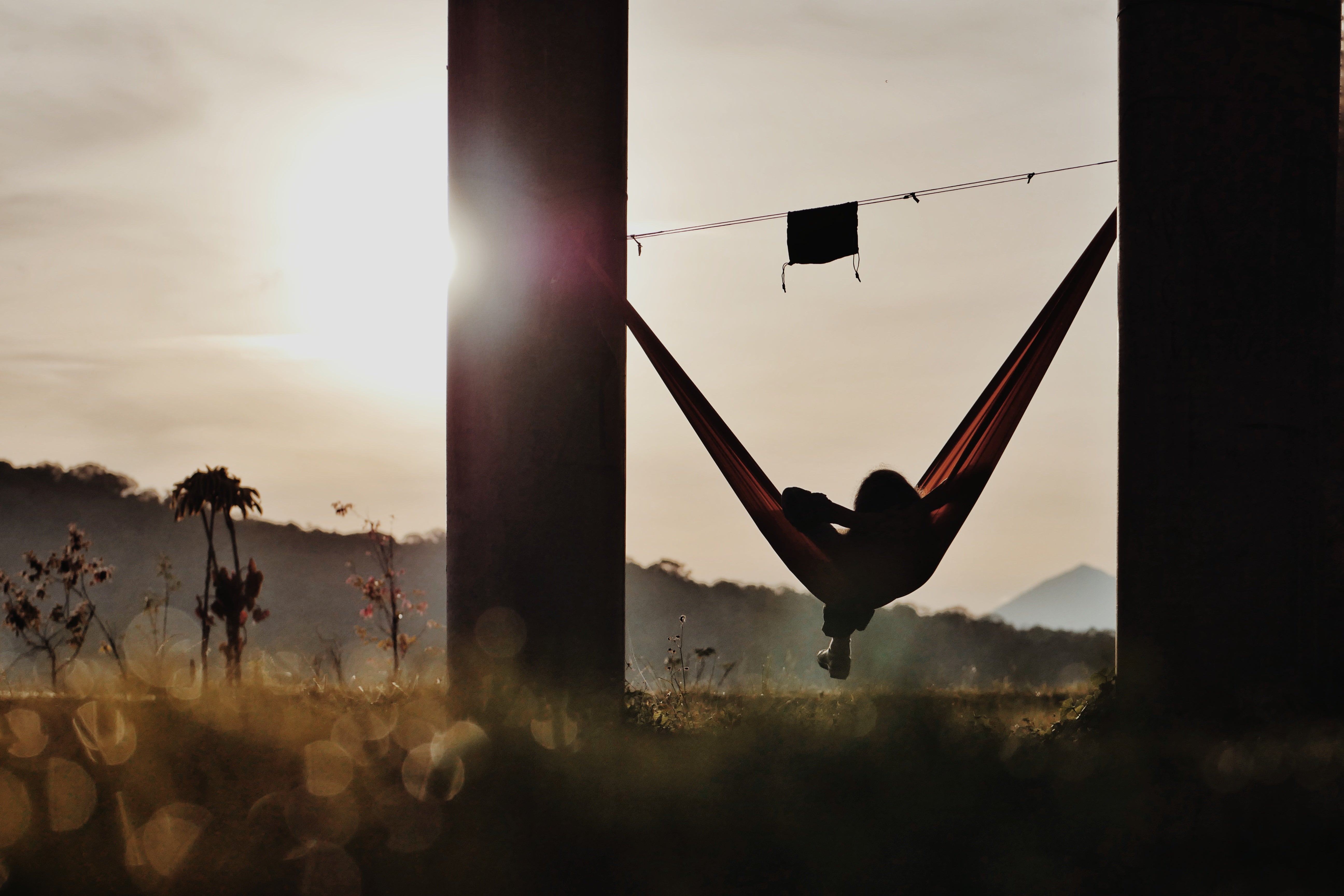 Person liegt in Hängematte bei Sonnenuntergang, Ridgeline mit befestigtem Netzfach gespannt zwischen Hängemattenenden, Silhouette vor Landschaft und untergehender Sonne