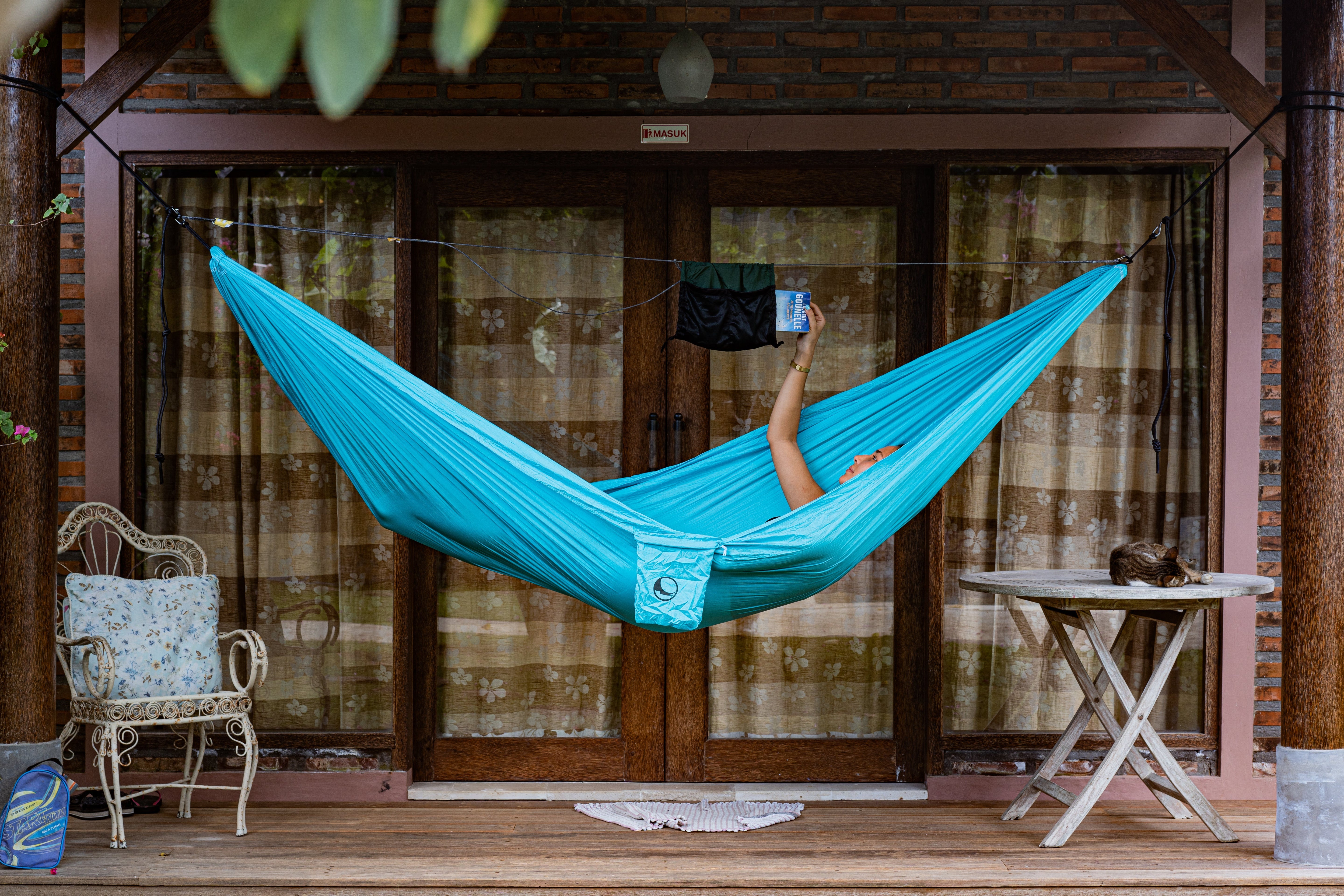 Person liegt in türkisfarbener Hängematte auf einer Veranda und greift mit der Hand zur schwarzen Ridgeline-Tasche, die an der Leine über der Hängematte befestigt ist