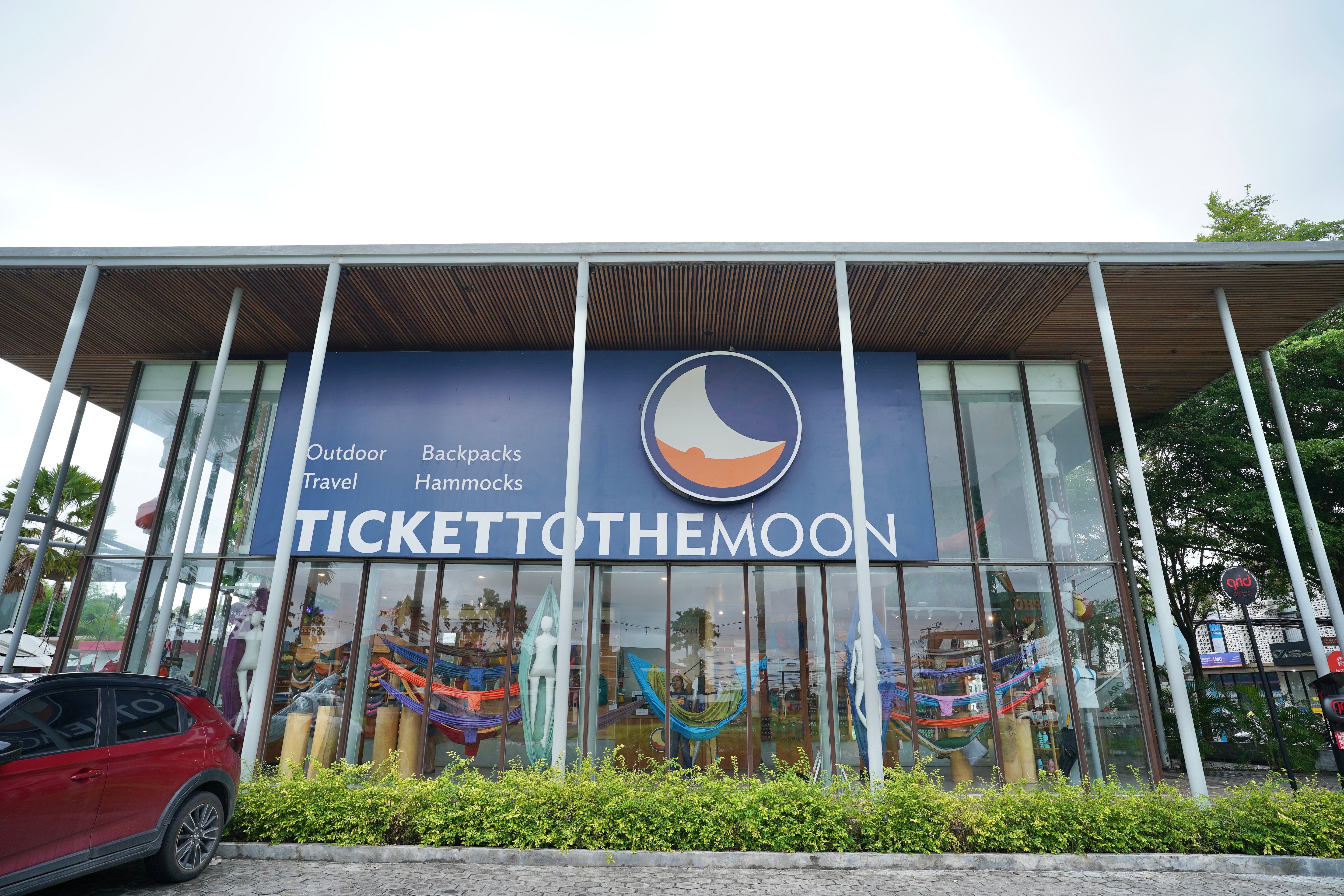 Moderne Glasfassade des Ticket to the Moon Stores in Bali/Berawa mit großem Logo, den Begriffen Outdoor, Travel, Backpacks, Hammocks und bunter Produktpräsentation im Inneren.