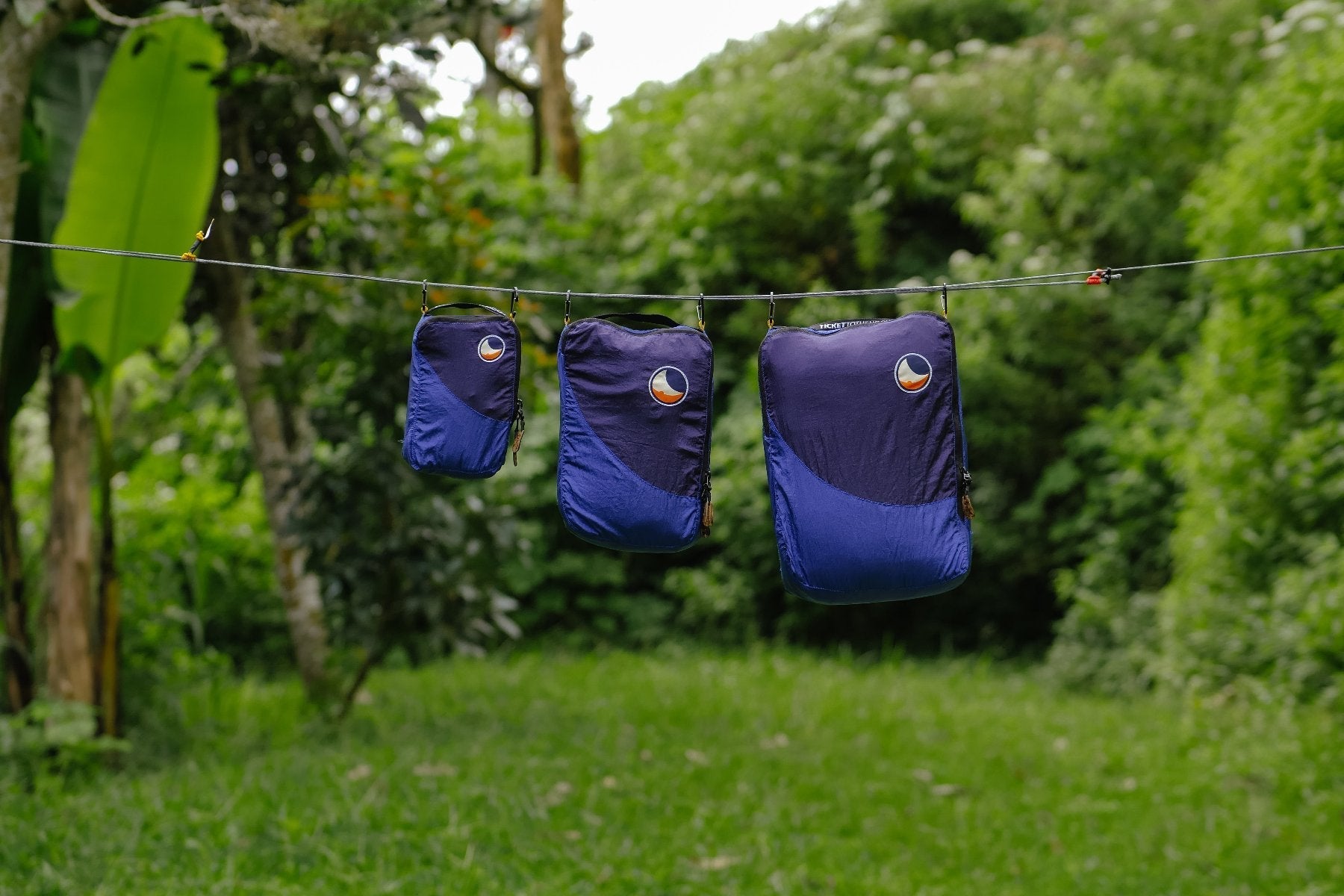Drei Travel Cubes in verschiedenen Größen hängen nebeneinander an einer Leine im Grünen – praktische Packwürfel für unterwegs.