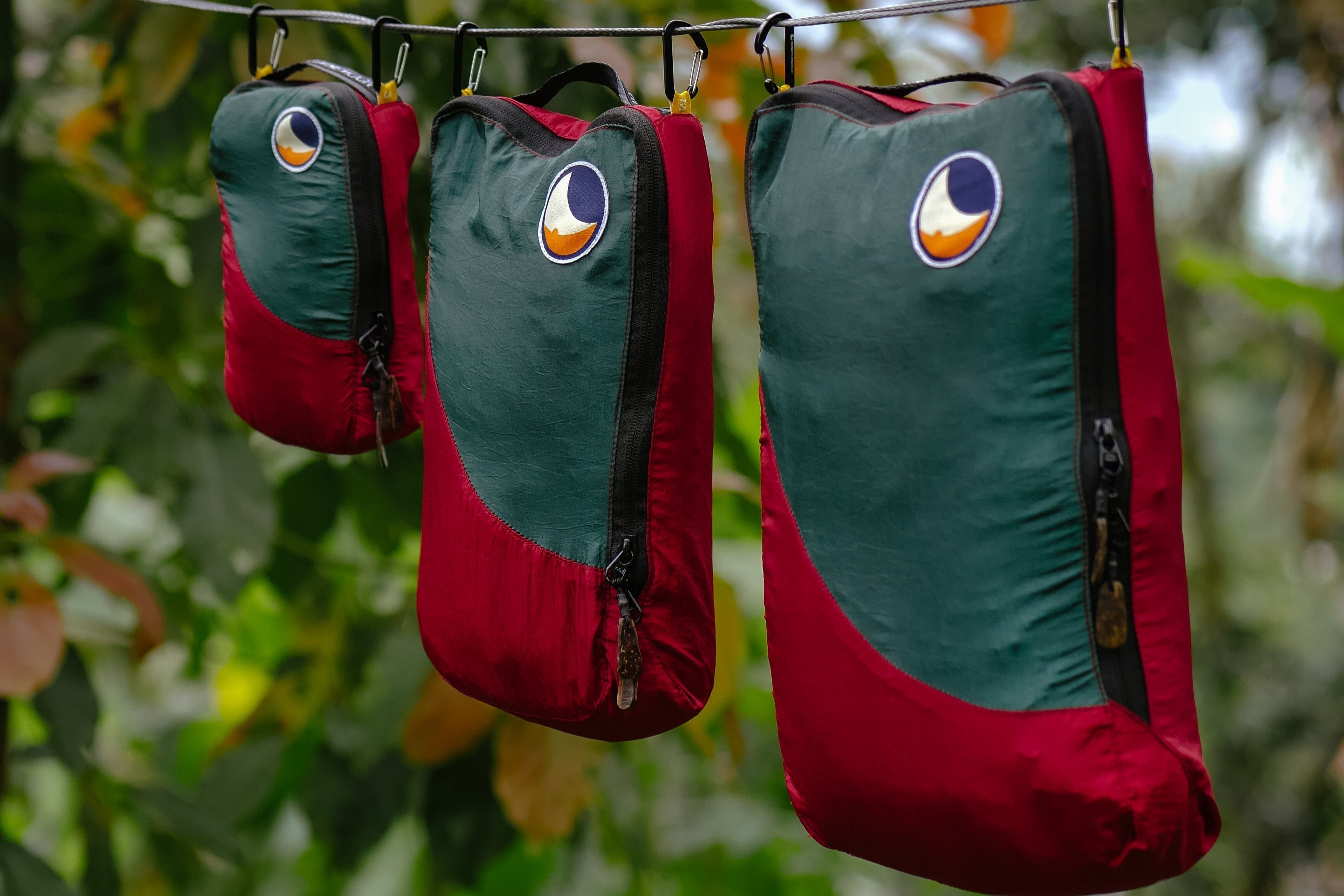 Drei rot-grüne Travel Cubes hängen an Haken vor einem grünen Waldhintergrund – stylisch und funktional für unterwegs.