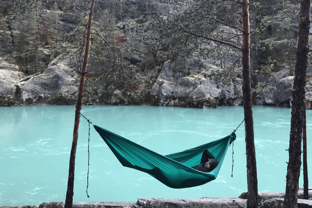 Grüne Hängematte an zwei Bäumen über einem türkisfarbenen Fluss, mit einer Person, die entspannt in der Hängematte liegt.