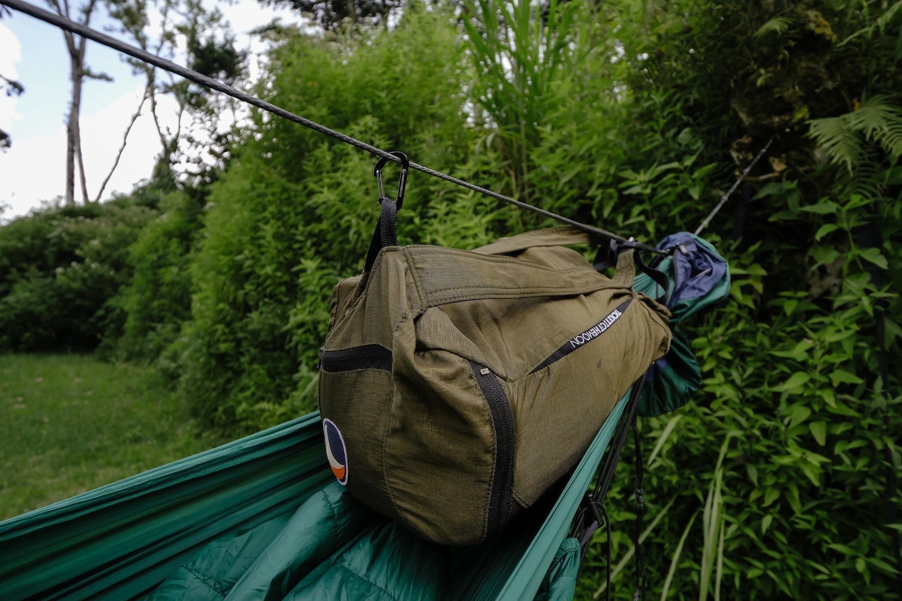 Kleine Tasche hängt mithilfe eines Accessory-Karabiners von Ticket to the Moon an einer Aufhängeschnur über einer Hängematte im Grünen.