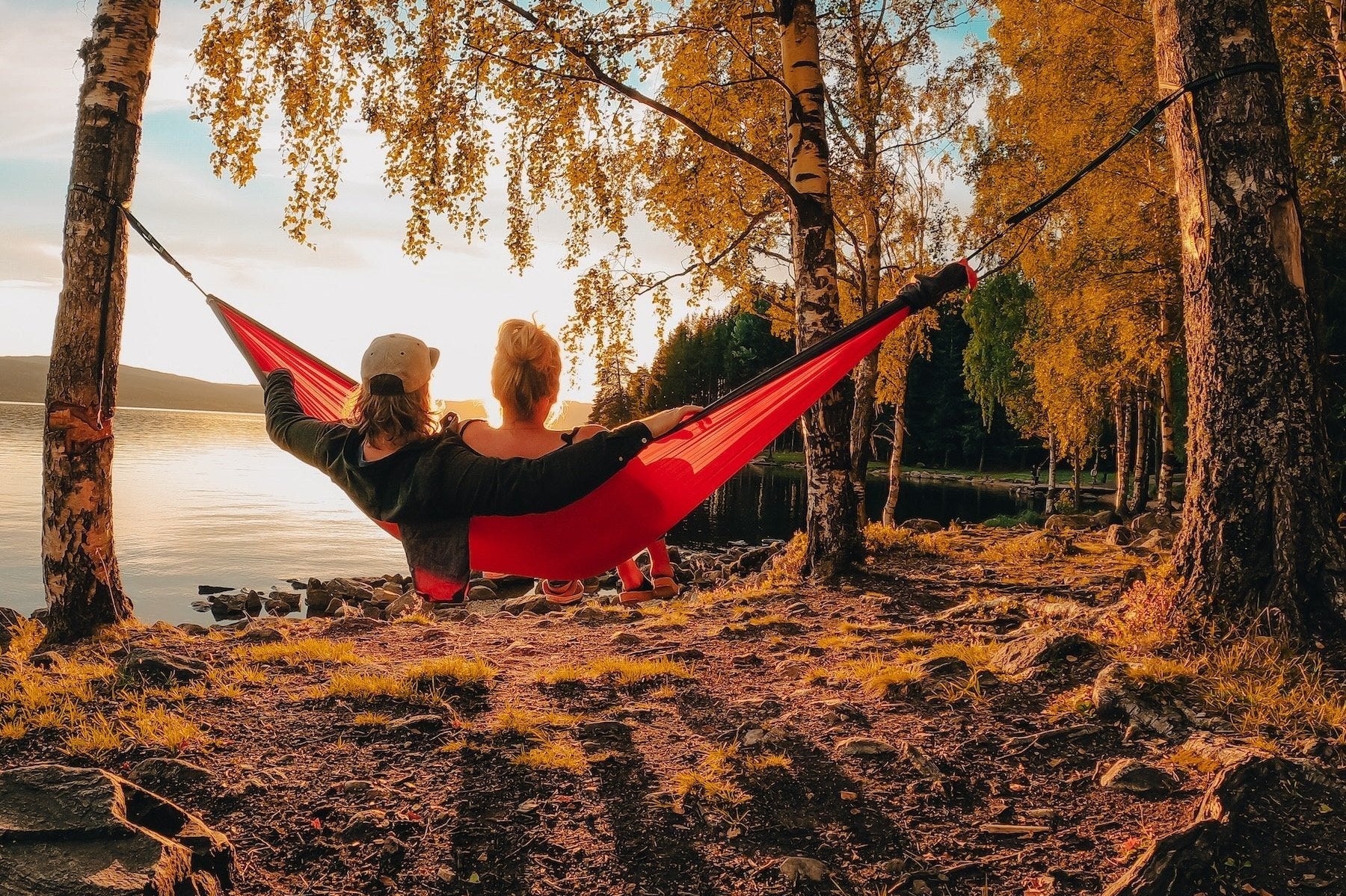 Pärchen entspannt in einer roten Ticket to the Moon Hängematte zwischen Birkenbäumen bei Sonnenuntergang am See