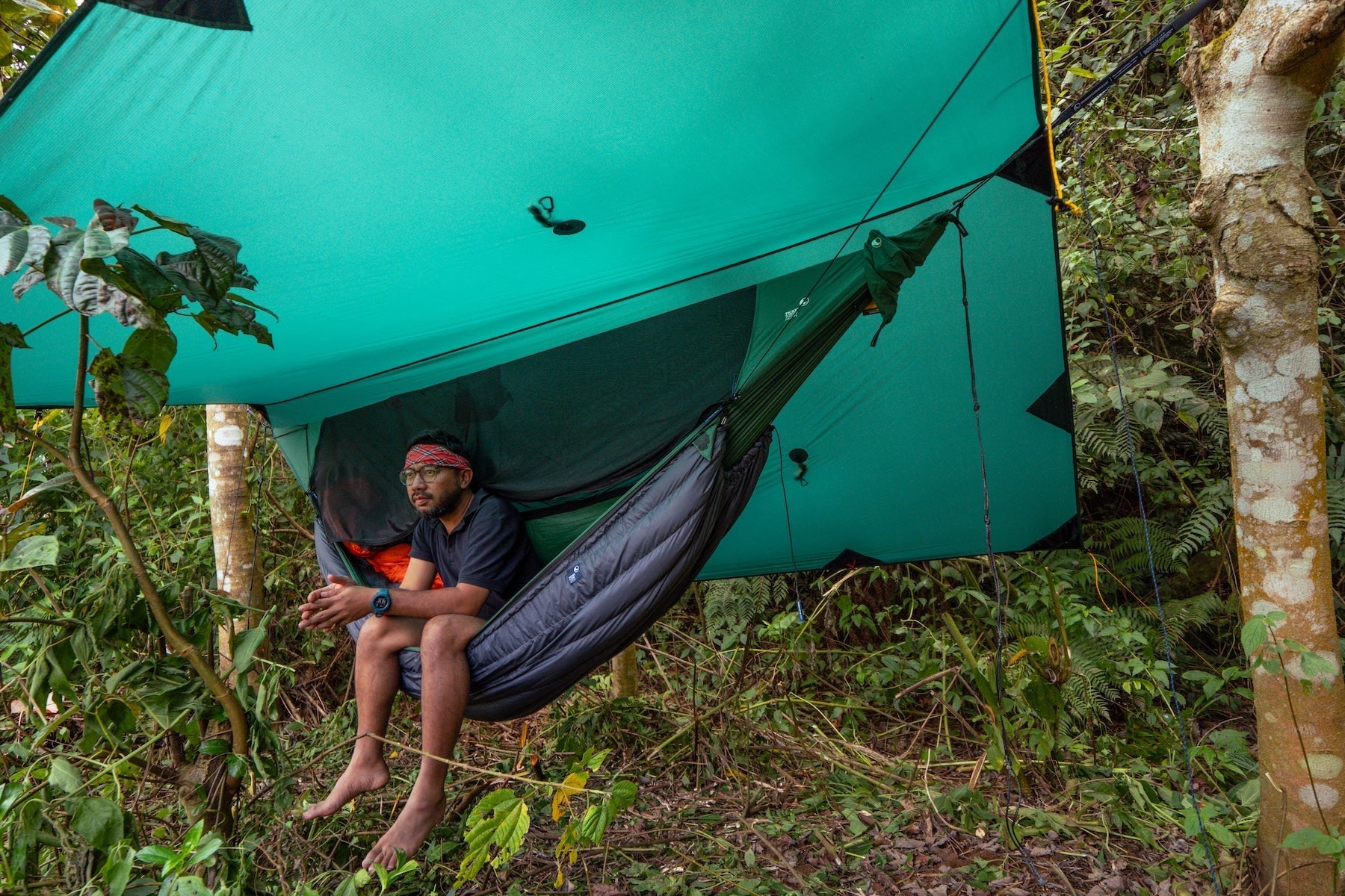 Mann sitzt in einer Ticket to the Moon Hängematte mit Underquilt unter einem Tarp inmitten des Dschungels