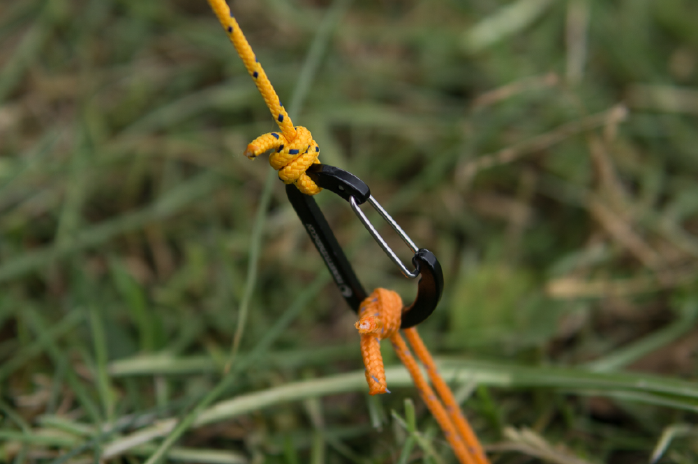 TTTM Karabiner bei der Abspannung eines Zelts oder Tarps auf Grasboden mit gelber und oranger Schnur