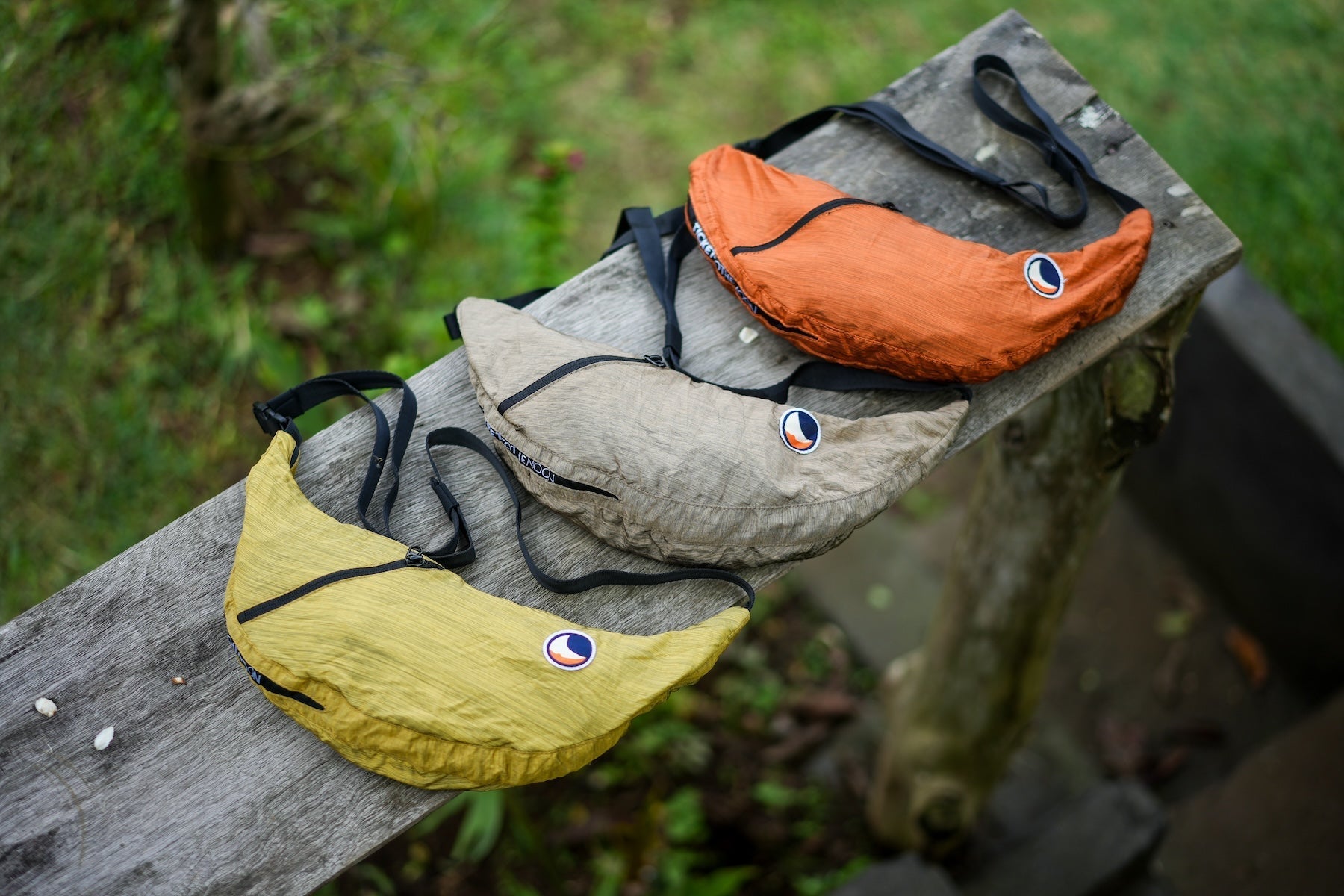 Drei TTTM Sling Bags in Erdtönen – Senfgelb, Sandbeige und Rostorange – liegen nebeneinander auf einer hölzernen Bank im Grünen