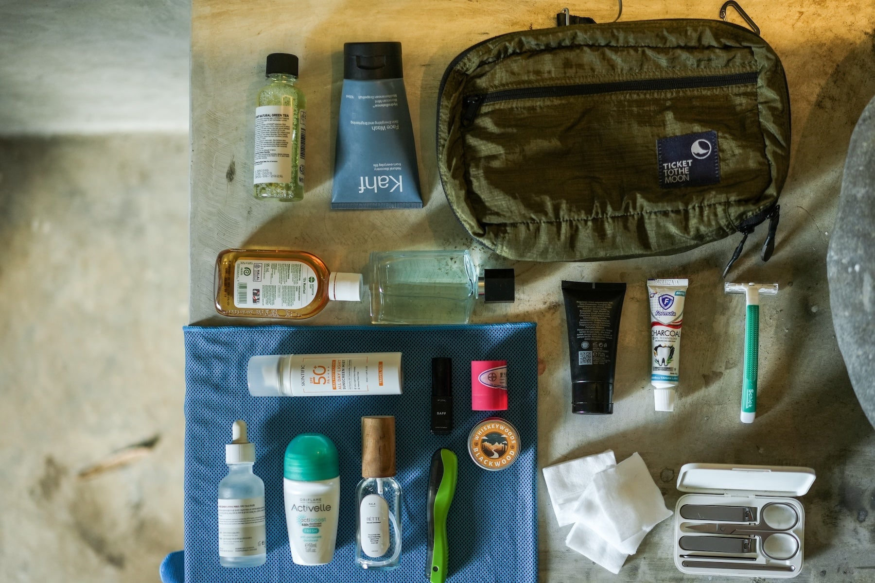 Washbag von Ticket to the Moon in Olivgrün im Flatlay-Stil, umgeben von verschiedenen Pflegeprodukten wie Zahnpasta, Sonnencreme, Shampoo, Nagelset und Rasierer – ideal für organisierte Reisen.