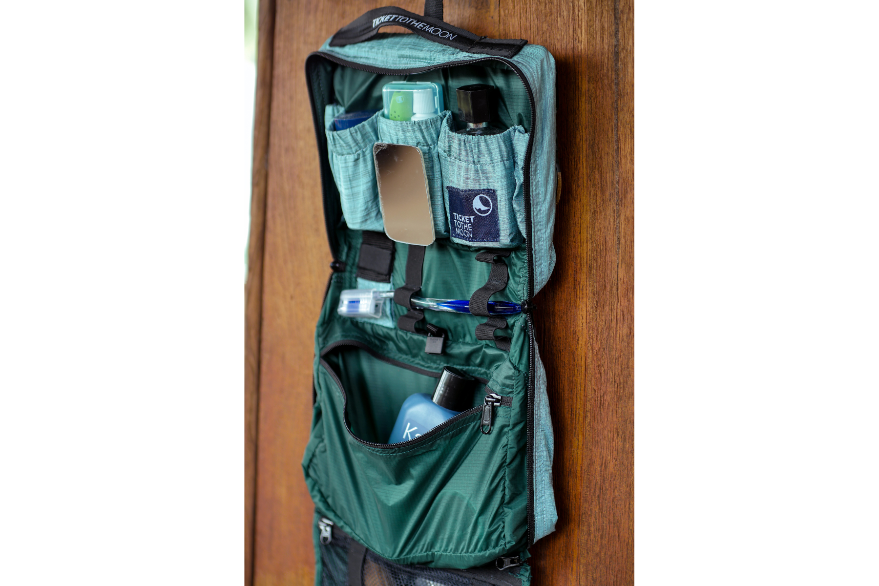 Geöffnete und befüllte türkise Washbag von Ticket to the Moon, an einer Holzwand aufgehängt – mit Spiegel, Fläschchen, Zahnbürstenetui und Hygieneartikeln übersichtlich verstaut.