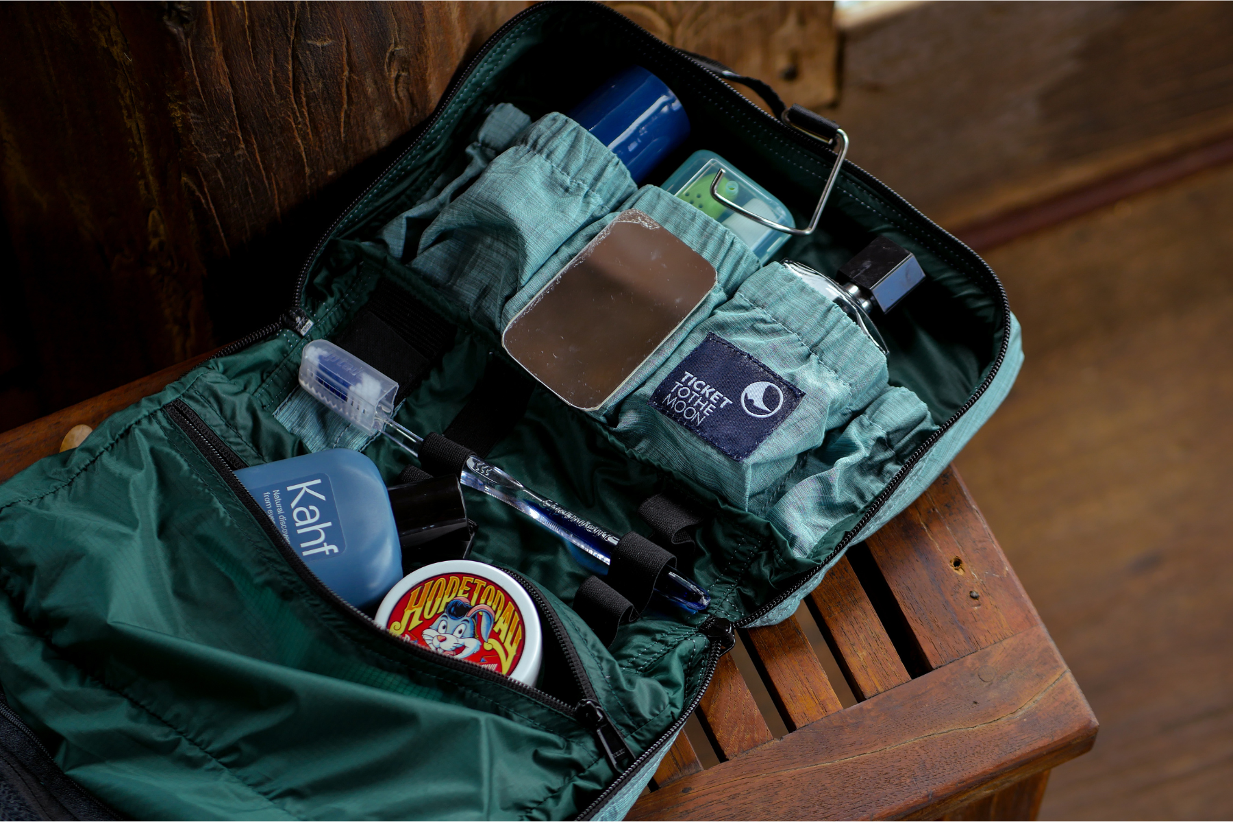 Nahaufnahme einer geöffneten, türkisfarbenen Washbag von Ticket to the Moon, liegend auf einer Holzbank – befüllt mit Pflegeprodukten, Spiegel und Zahnbürste für strukturierte Reiseorganisation.