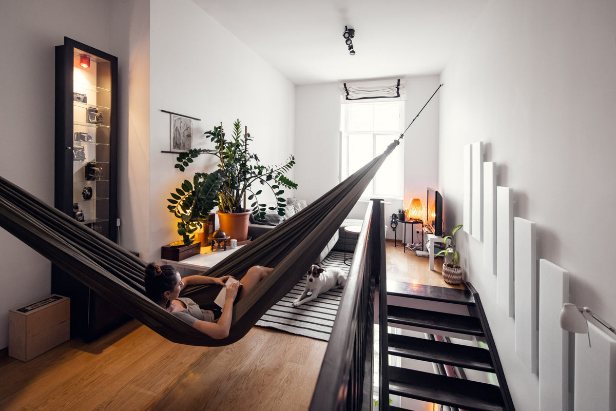 Frau liest in einer Home Hammock 320 in Olive Brown, quer durch eine helle Wohnung gespannt, darunter liegt ein Hund