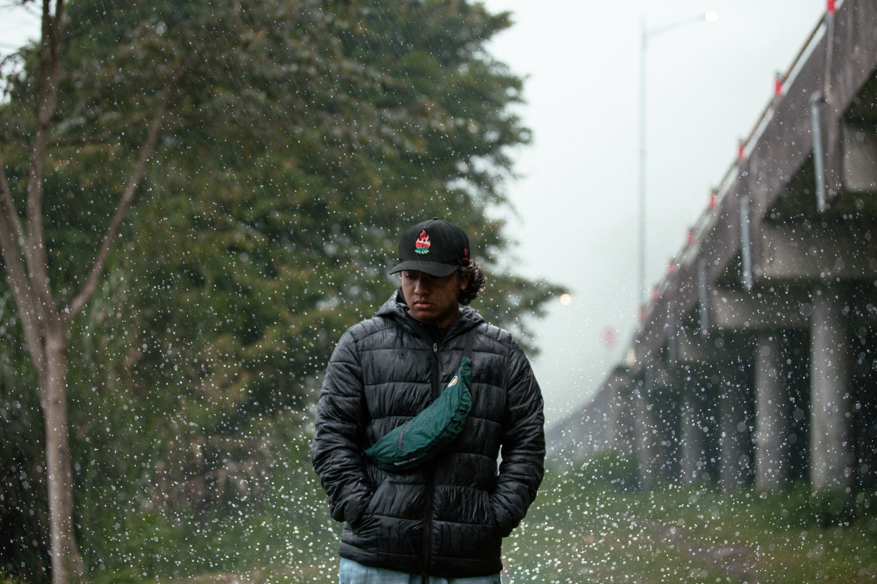 Ein Mann mit schwarzer Steppjacke und Basecap trägt die dunkelgrüne Water Resistant Ticket To The Moon Sling Bag bei starkem Regen im Freien um die Funktionalität der wetterfesten Umhängetasche aus Fallschirmnylon unter extremen Bedingungen zu demonstrieren.