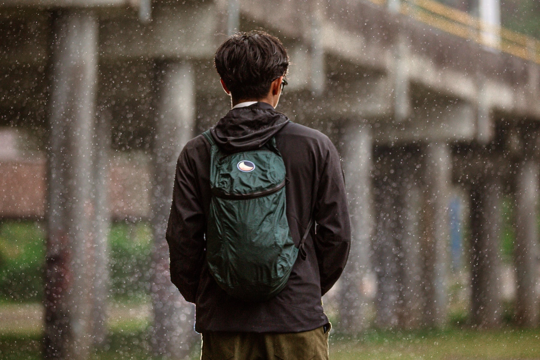 Rückansicht einer Person, die den dunkelgrünen Ticket to the Moon Mini Backpack während eines Regenschauers trägt. Das Bild verdeutlicht die wasserabweisende Funktion des Rucksacks in einer urbanen Outdoor-Umgebung.