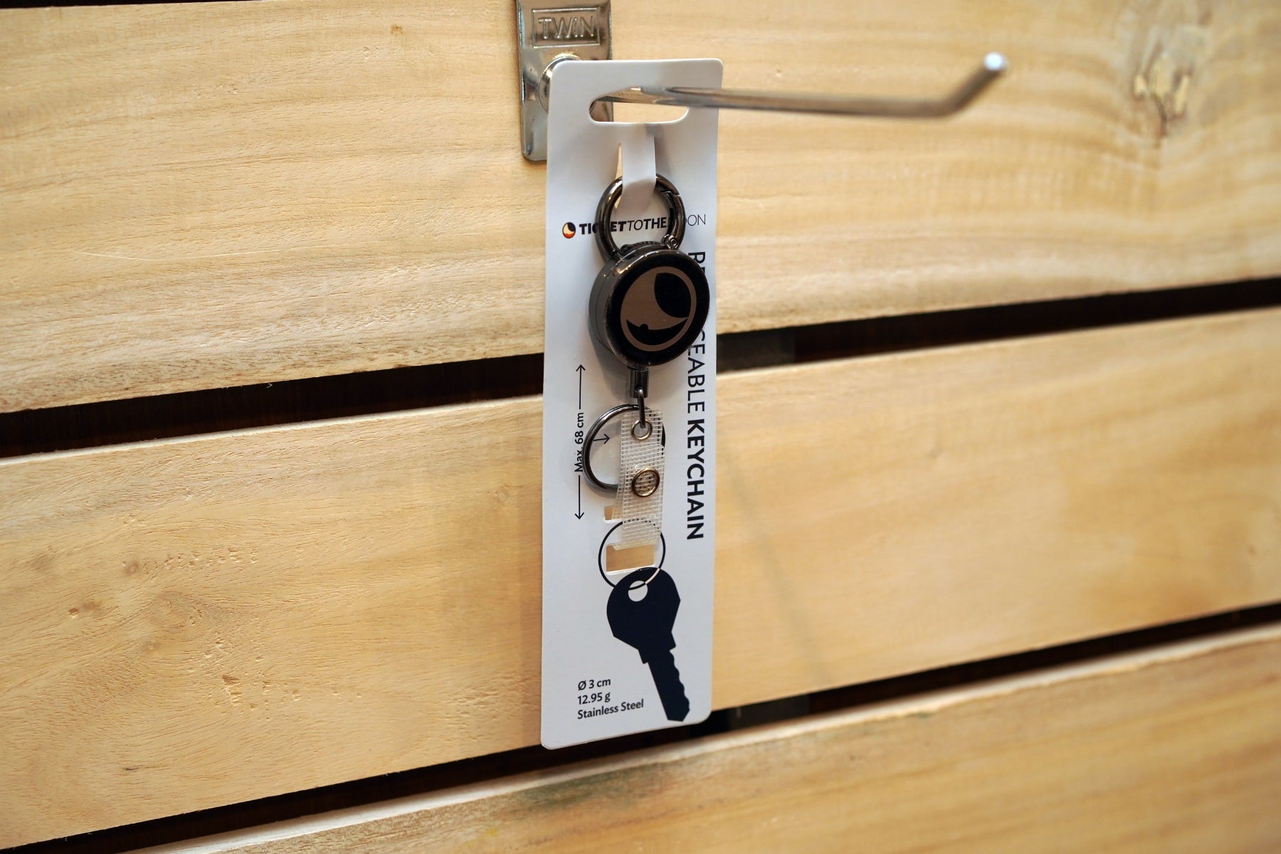 Der ausziehbare Schlüsselanhänger von Ticket to the Moon in seiner weißen Originalverpackung, hängend an einem Metallhaken vor einer rustikalen Holzwand.