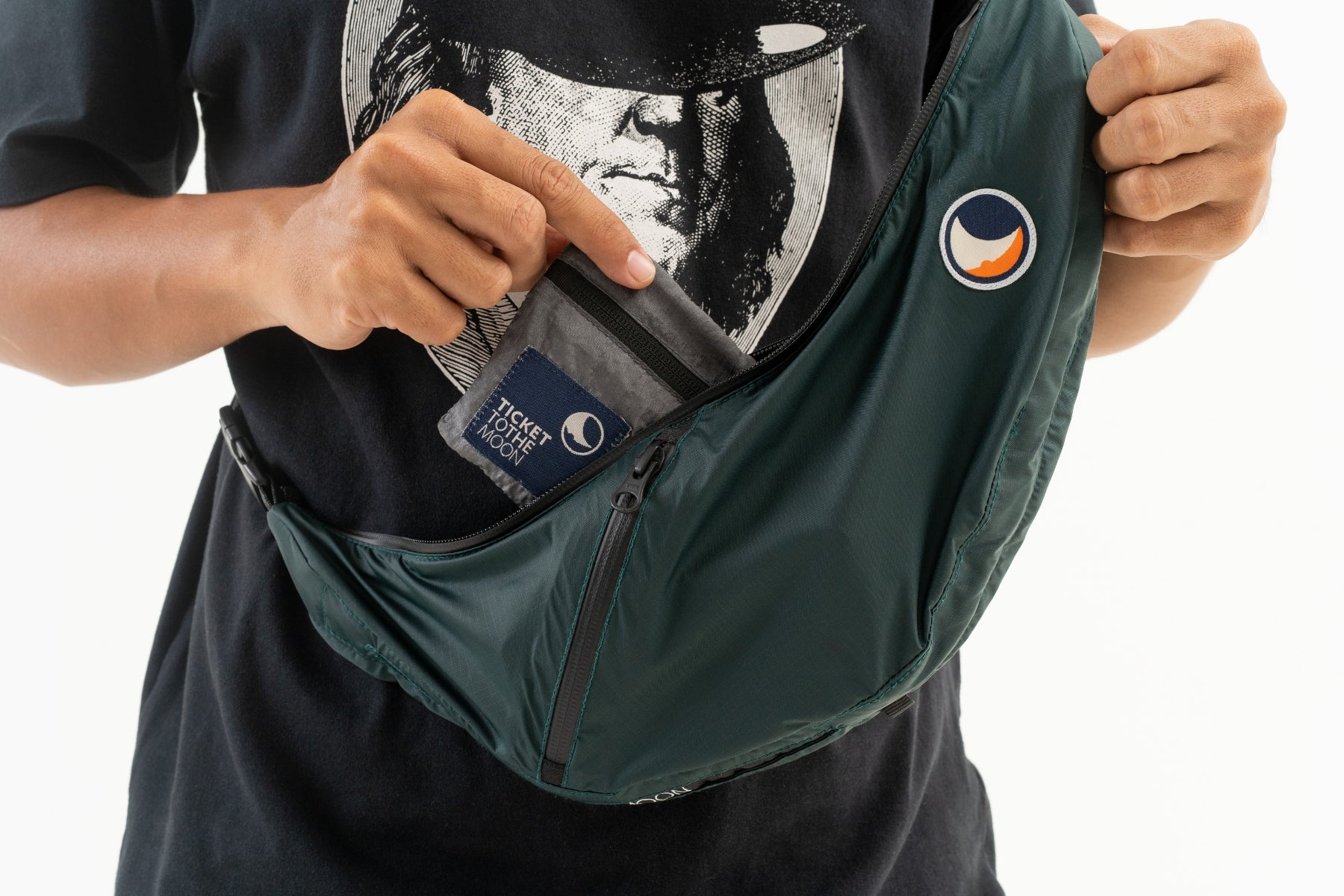 Nahaufnahme einer Person die eine dunkelgrüne wasserabweisende Ticket To The Moon Sling Bag trägt und ein graues Wallet in das Reißverschlussfach schiebt, wobei das markante Logo auf dem Fallschirmnylon sichtbar ist.