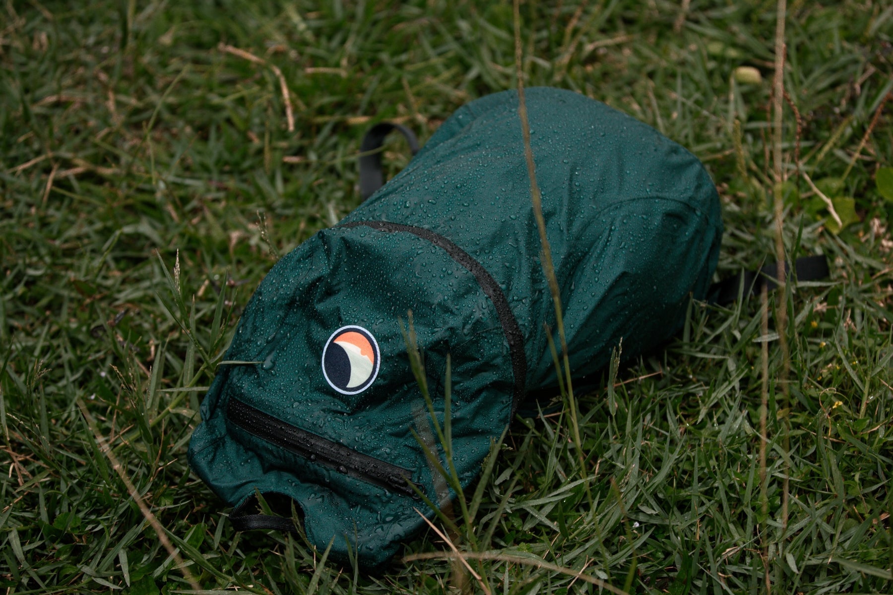 Der dunkelgrüne Ticket to the Moon Mini Backpack liegt im feuchten Gras. Wassertropfen perlen deutlich an der PU-beschichteten Oberfläche ab, was den Schutz vor leichtem Regen demonstriert.